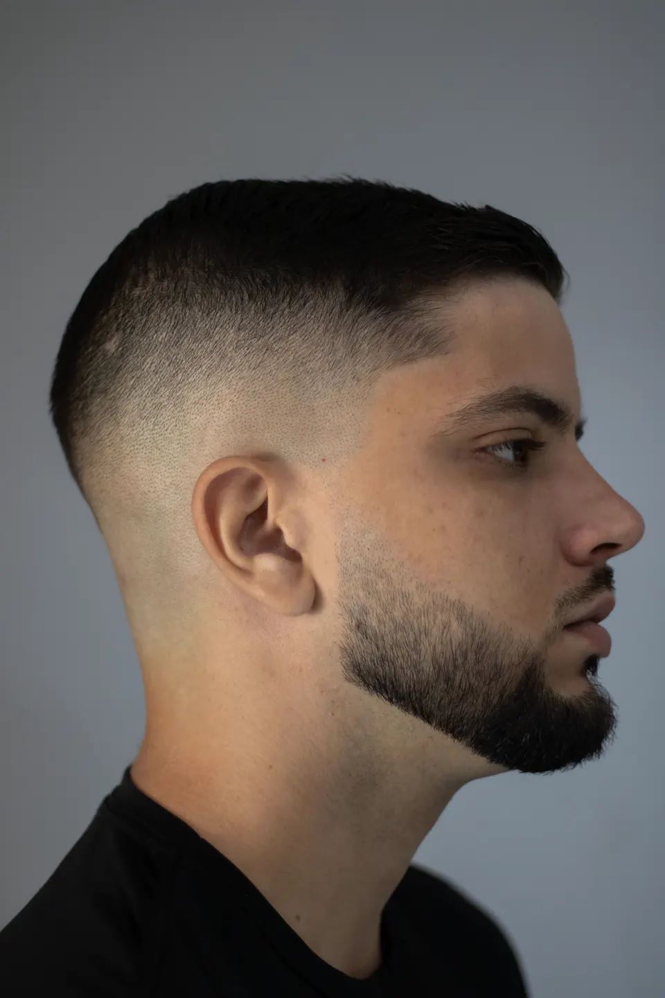 Profil eines Mannes mit sauberem Skin Fade und gepflegtem Kinnbart vor grauem Hintergrund