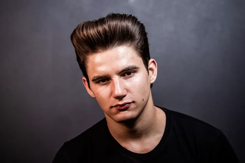 Porträt eines jungen Mannes mit hochgestyltem Pompadour vor dunklem Studiohintergrund